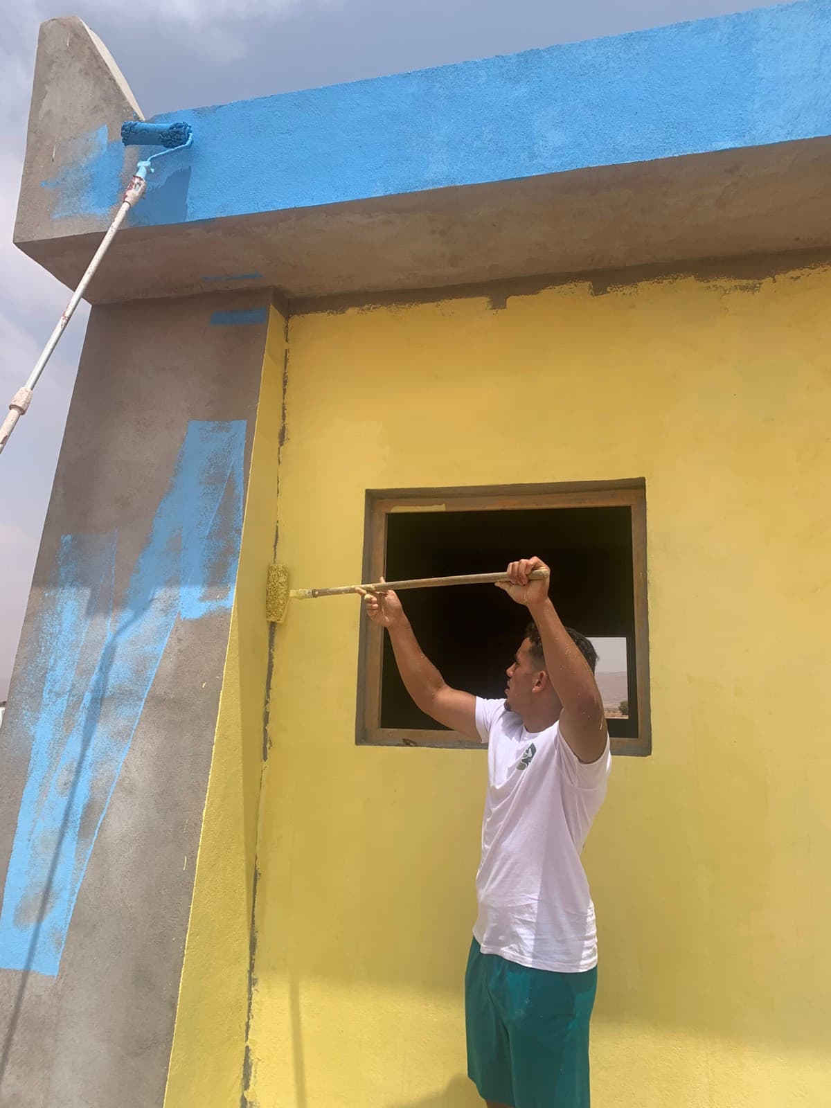 Un bénévole peint au rouleau le bandeau bleu qui couronne la façade jaune de l'école.