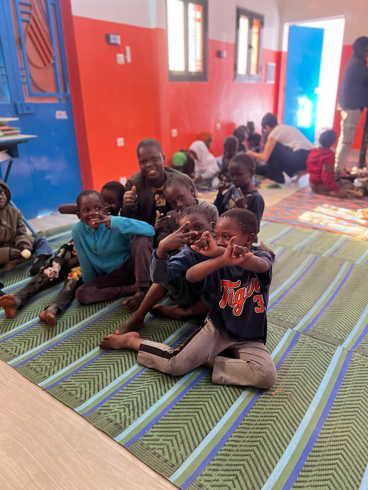 Des enfants assis sur une natte dans une salle aux murs bleus et orange sourient à l'objectif.