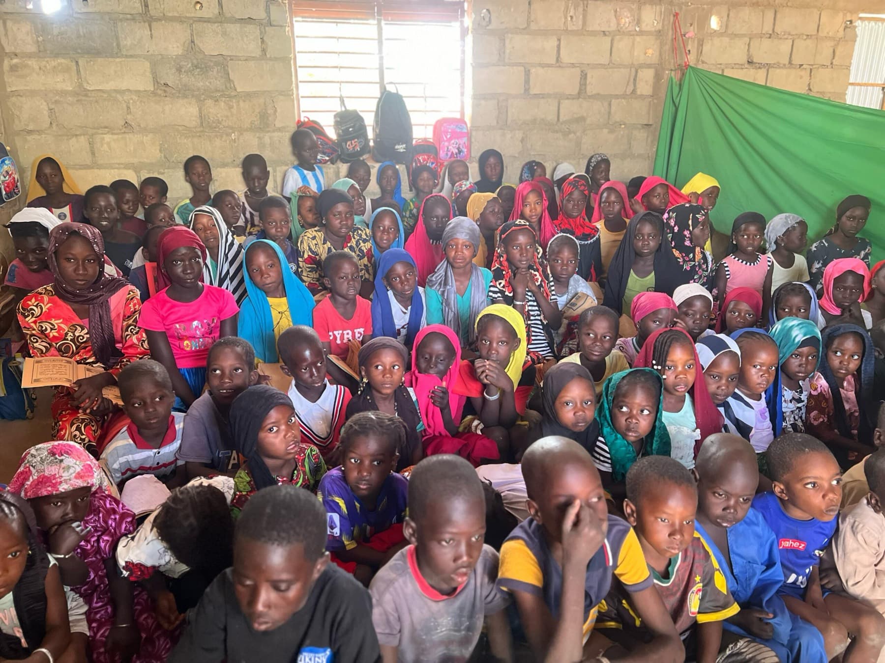 Des dizaines d'enfants assis serrés les uns contre les autres dans une salle aux murs de parpaings.