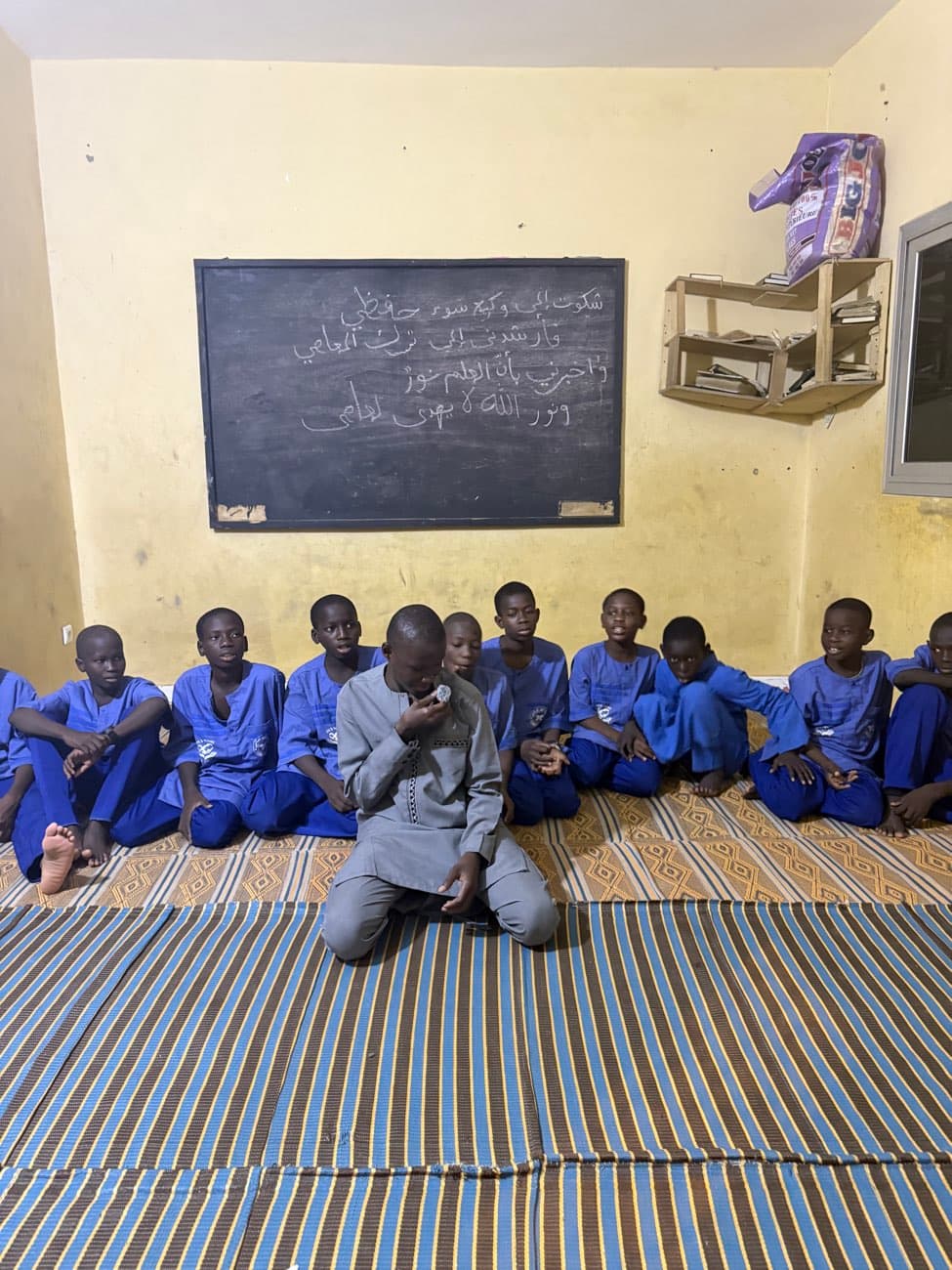 Des enfants en tenue bleue assis par terre dans une salle de classe, devant un tableau noir.