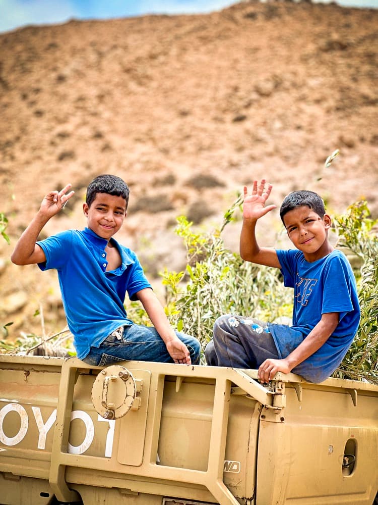 Deux enfants assis à l'arrière d'un pick-up saluent la caméra.